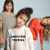 Stylish Bone Colored Crewneck Sweatshirt Worn by Adorable Young Girl. Logo in Velvet Lettering. 100% Organic Cotton. Smaller Things Logo Crewneck for Young Boys and Girls.